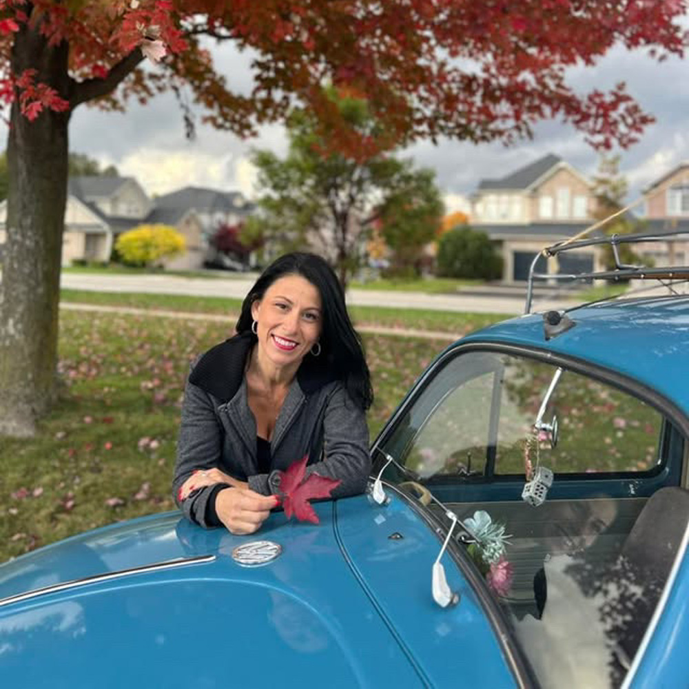 Sandra holding red autumn leaf while leaning on blue VW Beetle under fall foliage