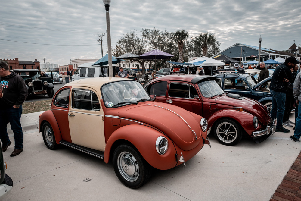 Two tone Volkswagen Beetles parked at a 2026 VW meet with crowds and tents in the background