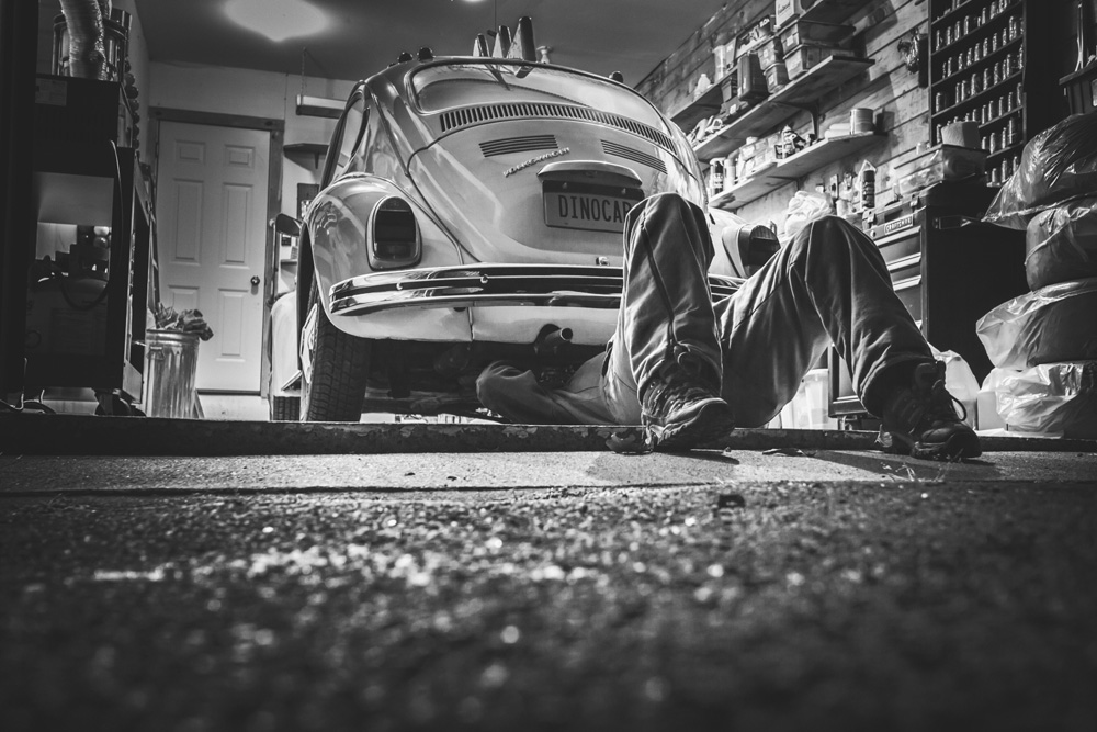 Person working under classic Volkswagen Beetle in garage with tools and supplies on shelves