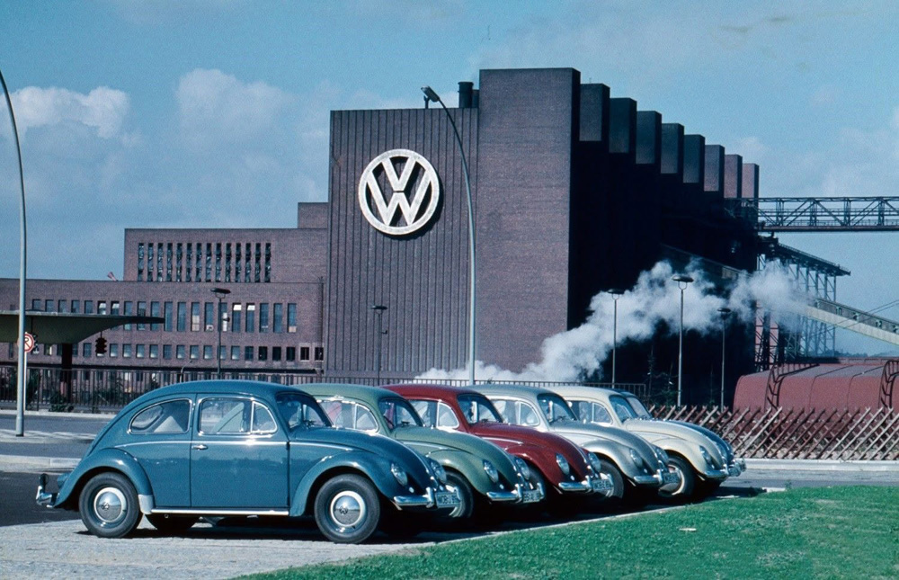 Colourful late-1950s Volkswagens parked near Wolfsburg station with the VW factory Heizkraftwerk power plant in the background